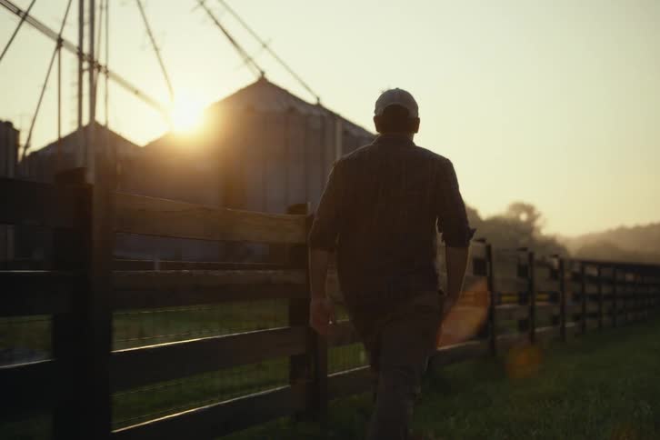 A farmer walks along a country fence toward his grain silos as the sun rises over the horizon