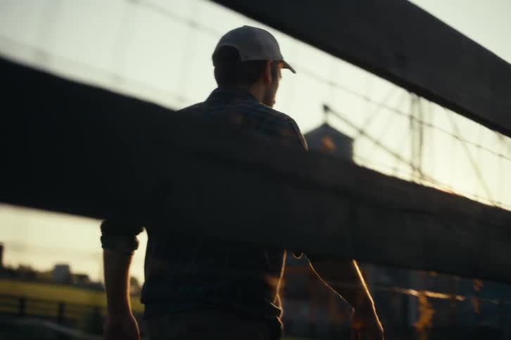 Viewed from behind a wooden fence, a man in a plaid shirt and hat walks across a rural farm during a golden sunset