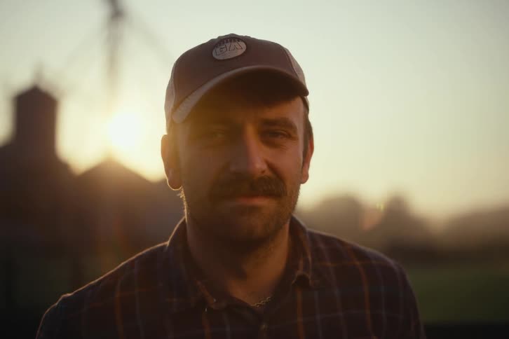Portrait of a proud farmer with a mustache looking into the camera with a calm expression, backlit by the golden hour sun on his farm
