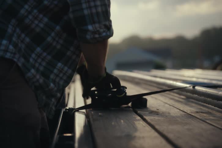 A blue-collar worker in a plaid shirt and gloves tightens a ratchet strap on a flatbed trailer at dawn