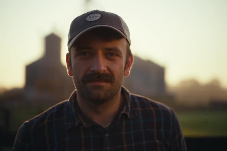 Portrait of a mustachioed farmer in a hat who looks up and winks at the camera during a beautiful golden hour sunset