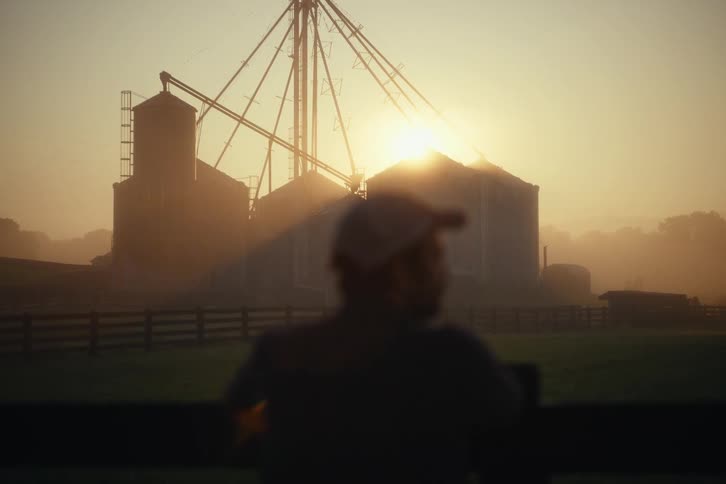 A farmer in silhouette looks out over his silos and fields during a beautiful hazy sunrise