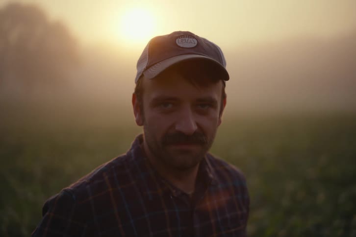 A thoughtful farmer in a plaid shirt and baseball cap looks directly at the camera in a foggy field at sunrise