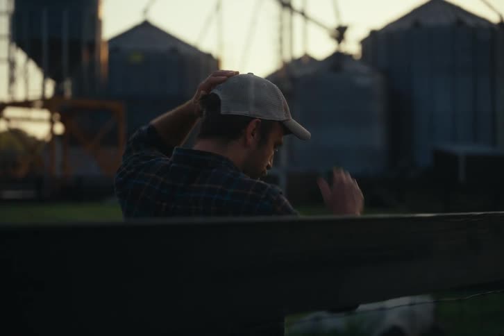 A farmer seen from behind adjusts his cap while looking out at grain silos during a golden sunset