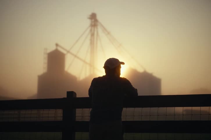 A silhouetted farmer wearing a hat leans on a fence, watching the sunrise over misty farm silos