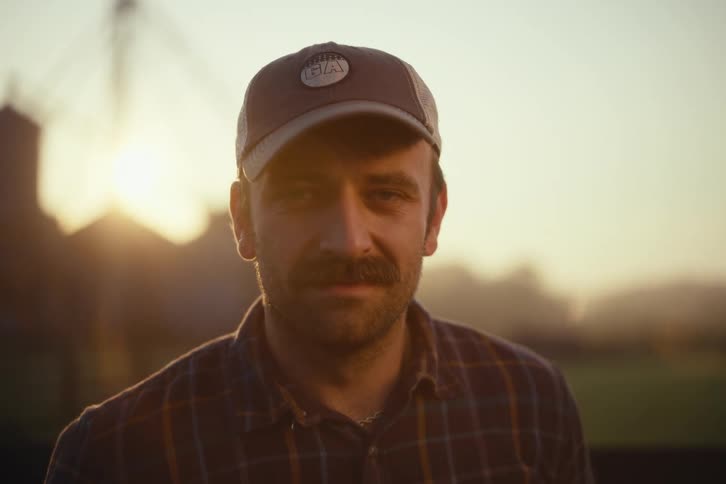 Close-up portrait of a serious farmer with a mustache looking at the camera during a golden hour sunset on his farm