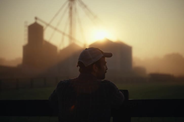 A lone farmer surveys his land and silos, silhouetted by the golden light of a misty sunrise