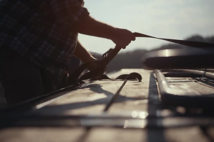 Backlit by the bright sun, a farmer in a plaid shirt secures a metal gate to a flatbed trailer with a ratchet strap