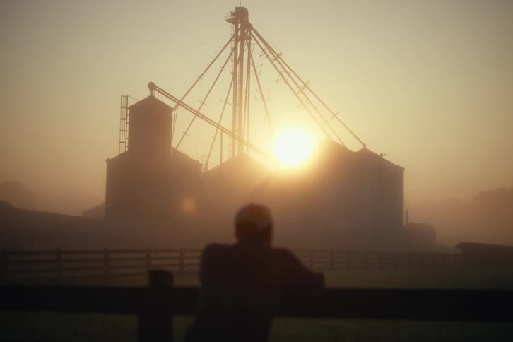 A farmer leans on a fence looking out at grain silos as the sun rises on a beautiful, foggy morning