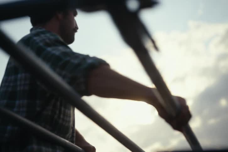 A farmer in a plaid shirt works with metal pipes on a trailer, silhouetted against the warm glow of a rural sunset