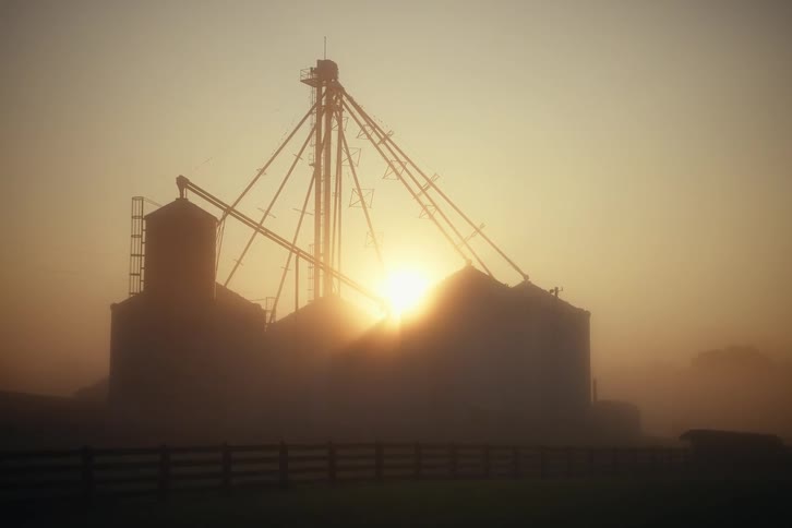 A rack focus reveals a silhouetted grain silo on a farm during a foggy, golden sunrise