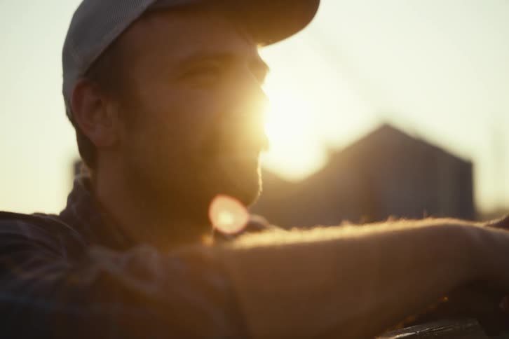 A thoughtful farmer with a mustache adjusts his cap, backlit by the setting sun with farm silos in the background
