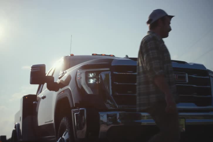 A determined man in a plaid shirt walks past his heavy-duty GMC truck as the bright sun shines down