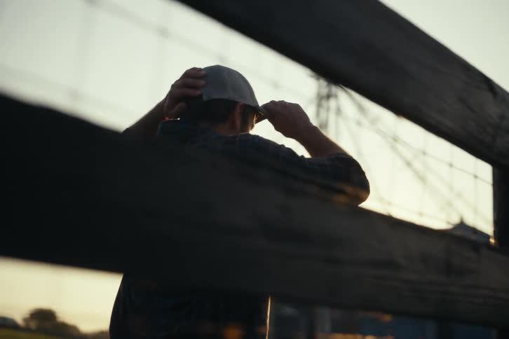 A farmer walks along a wooden fence line on his property, pausing to adjust his cap as the sun sets in the background