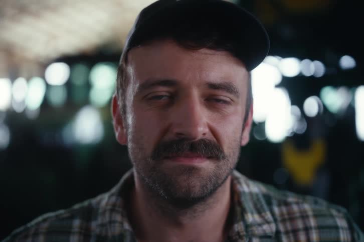 A hardworking man with a mustache and a plaid shirt lifts his head and stares intensely into the camera in a dimly lit workshop