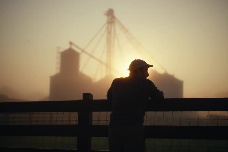 A silhouetted farmer leans on a fence, contemplating the misty sunrise over his farm's grain silos before walking off to start his day