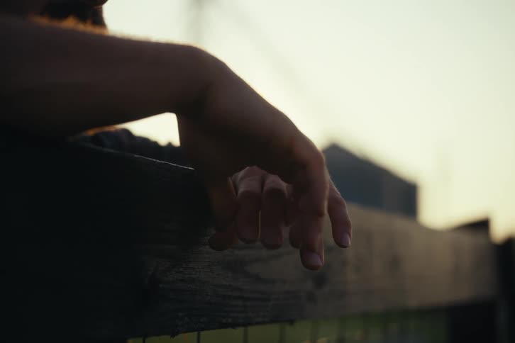 A person's hand rests on a wooden fence as the setting sun peeks through their arm, creating a beautiful lens flare