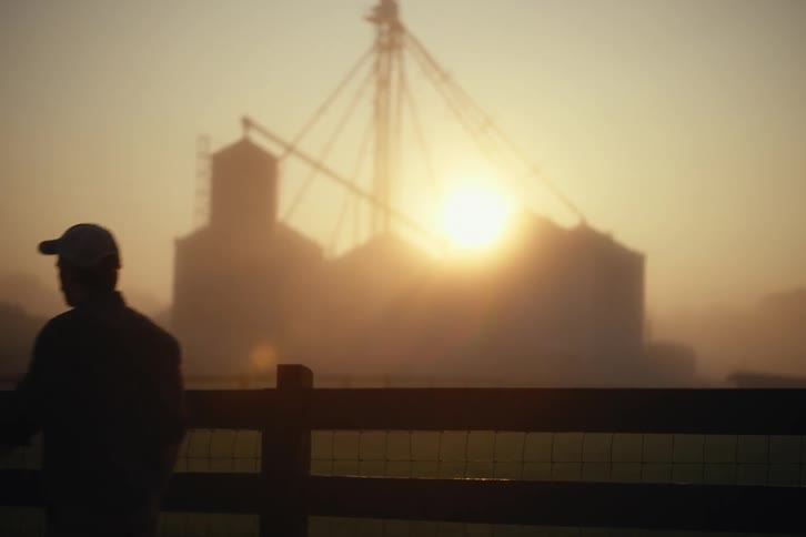 A farmer in silhouette walks across a field on a foggy morning as the sun rises behind large grain silos