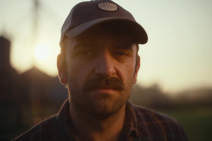 A cinematic close-up of a serious farmer with a mustache looking into the camera at golden hour with his farm in the background