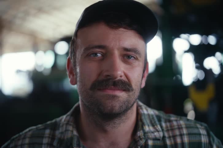 A close-up portrait of a confident man with a mustache wearing a plaid shirt and cap, looking calmly at the camera inside a rustic barn