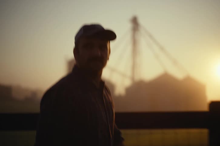 A thoughtful farmer adjusts his hat while walking along a fence at sunrise, with silos silhouetted against the golden sky
