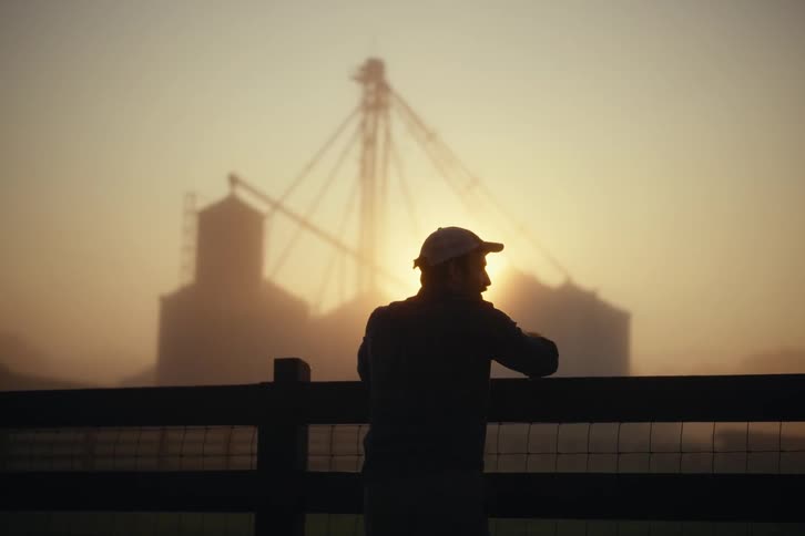 A silhouetted farmer leans on a fence and surveys his land at dawn with grain silos visible through the fog in the background