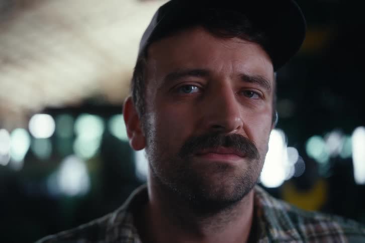 A close-up portrait of a serious man with a mustache and cap who offers a subtle smile while looking at the camera