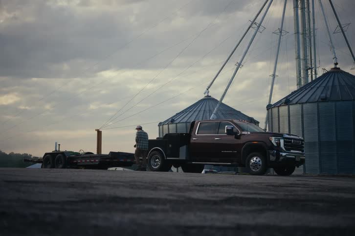 A farmer gets out of his dually truck with a flatbed trailer attached, standing in front of large grain silos under a cloudy sky