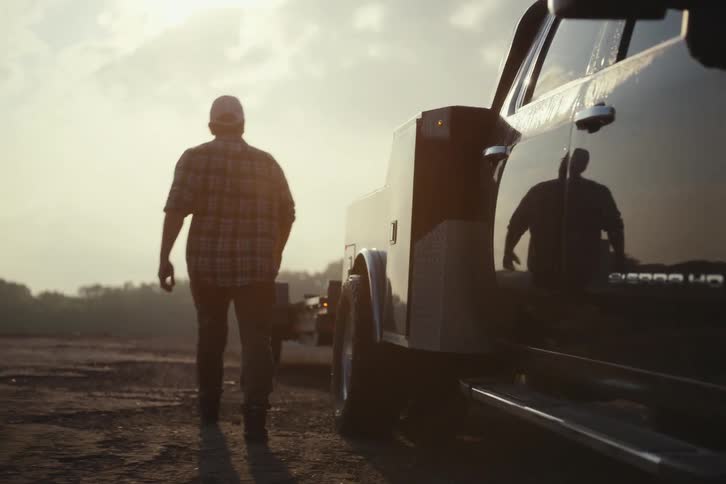 A man in a plaid shirt gets out of his heavy-duty pickup truck and walks toward the attached trailer in slow motion at sunrise