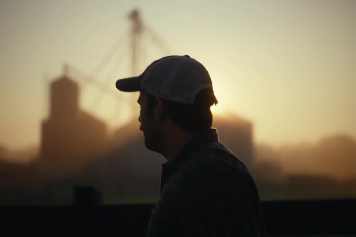 A silhouetted farmer in a hat looks out over his farm during a hazy sunrise, running his hands through his hair in slow motion