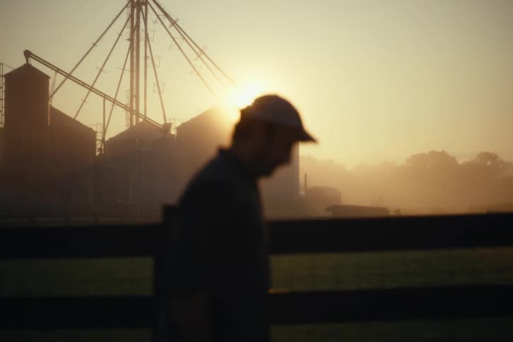 The silhouette of a farmer in a cap walks slowly past grain silos during a hazy golden sunrise