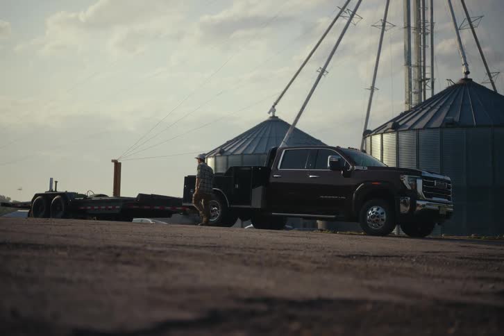 A male farmer in a plaid shirt gets out of his dually work truck with a flatbed trailer attached and walks to the back on a rural farm at sunset