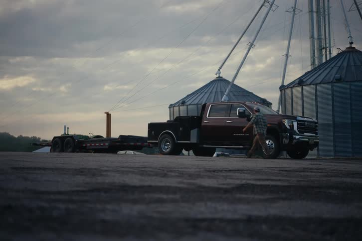 A man gets out of his large dually pickup truck with a trailer and walks around it on a farm with grain silos in the background