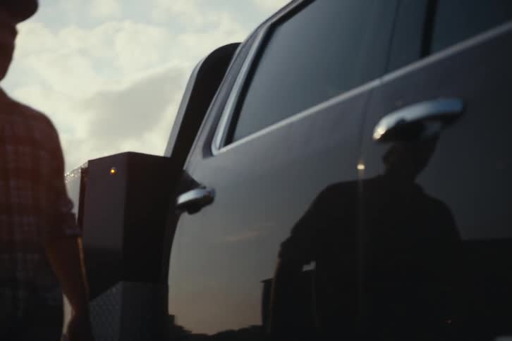 A serious man in a plaid shirt and hat walks away from his large truck in slow motion against a cloudy, sunlit sky