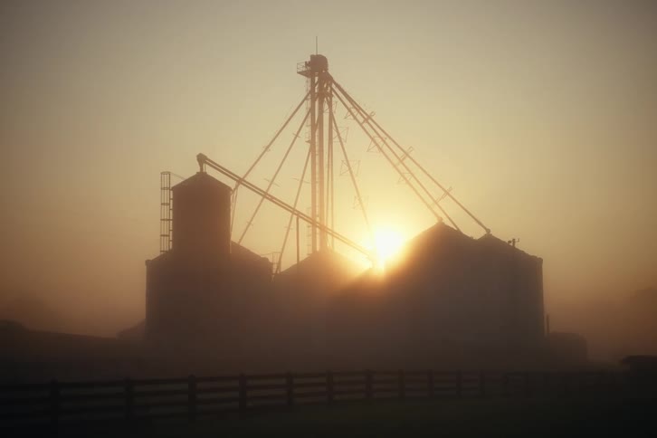 The silhouette of a large farm granary stands against the golden light of a foggy sunrise