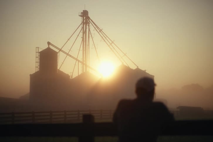 A silhouetted farmer stands at a fence and looks out at large grain elevators during a hazy golden sunrise