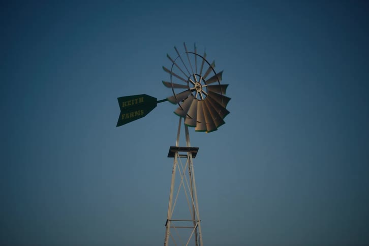 A classic metal farm windmill with 'Keith Farms' on its tail slowly turns against a clear blue sky