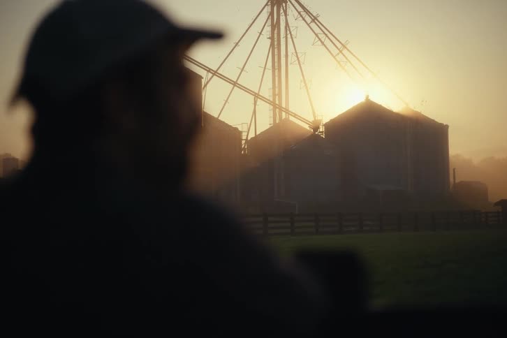 A silhouetted farmer tips his cap while looking out at a large grain silo complex during a hazy golden sunrise