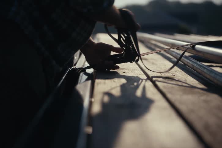 A silhouetted man fastens a ratchet strap to secure metal pipes onto a wooden flatbed trailer in the early morning light