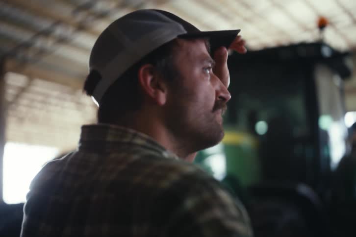 A hardworking farmer wearing a plaid shirt and hat wipes sweat from his brow while standing in his barn