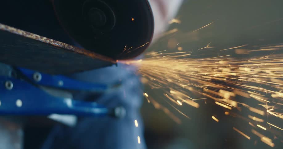 A close-up slow-motion shot shows an angle grinder cutting through a sheet of metal, sending bright sparks flying
