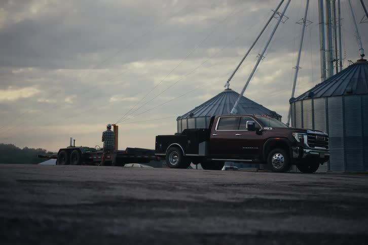 A farmer steps out of his heavy-duty pickup truck to check his trailer in front of grain silos on an overcast day