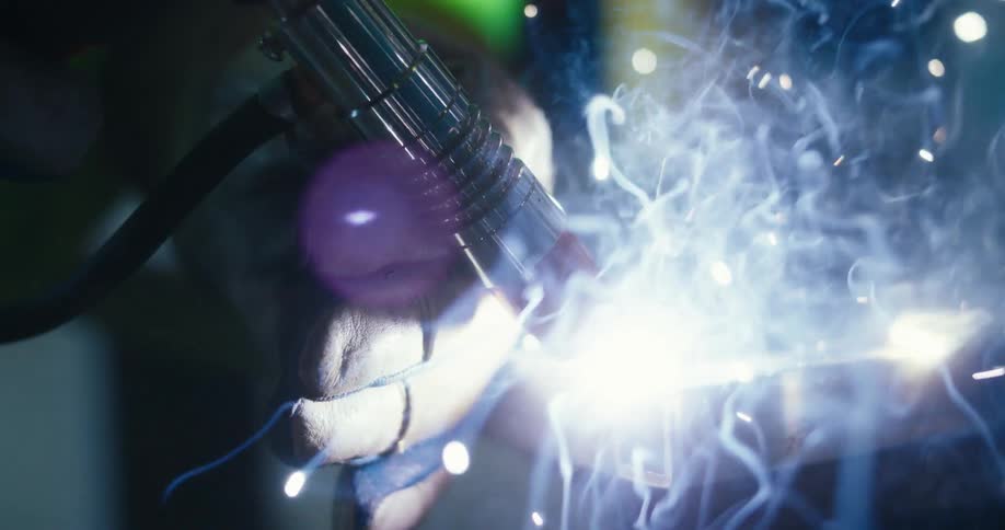 A close-up slow-motion shot captures a welder's gloved hand using a torch to fuse metal, creating a brilliant display of sparks and smoke