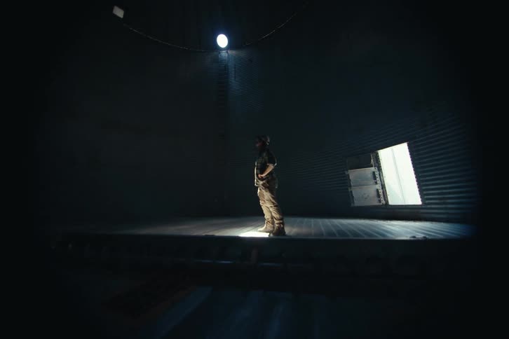 A farmer stands alone in a dramatic spotlight inside a dark and empty grain silo, inspecting the interior
