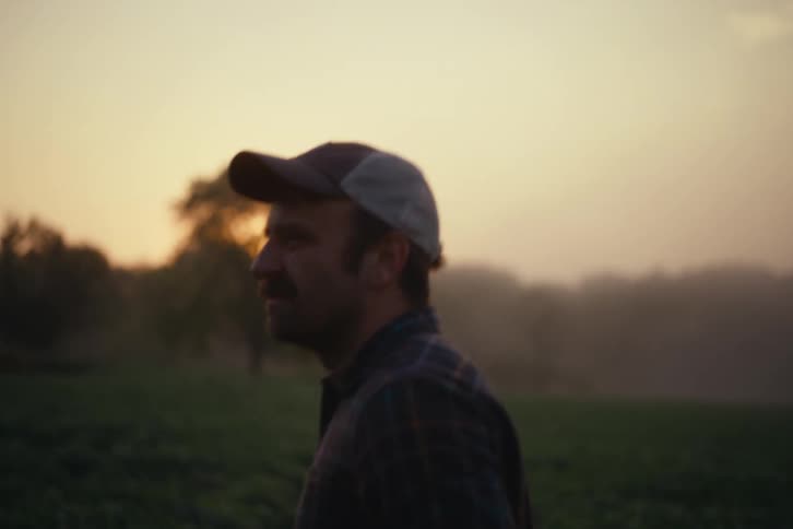 A farmer wearing a plaid shirt and cap walks through his field during a beautiful golden sunset