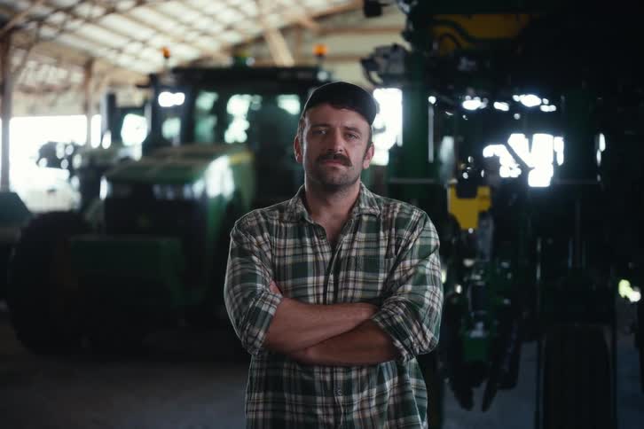 A proud and determined farmer with a mustache stands with his arms crossed in front of his tractors inside a large barn