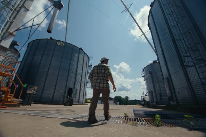 Slow motion low angle follow shot of a farmer walking between towering grain silos on a sunny day at a rural farm