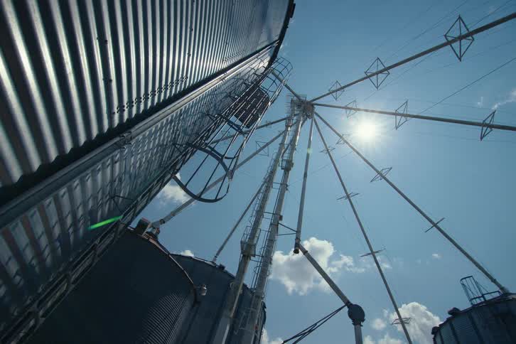 A wide, low-angle shot looks up at massive industrial grain silos against a bright blue sky with the sun shining and clouds passing by