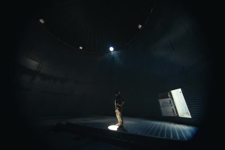 A slow tilt down reveals a lone man standing in a single beam of light inside a massive, dark, and empty industrial silo