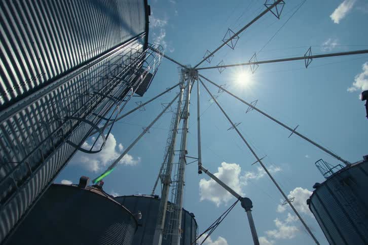A low-angle view of a towering grain silo complex against a bright sun and blue sky